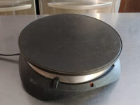An electric crepe maker with a round black cooking plate sits unused on a stainless steel counter, the appliance used to prepare Crepes.