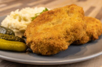 Two golden fried Dutch Schnitzel (Holandský Řízek) cutlets on a grey plate with gherkins and a shredded white side.