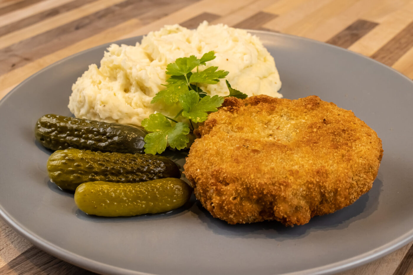 Golden fried Dutch Schnitzel (Holandský Řízek) served with mashed potatoes, pickles, and parsley on a gray plate.