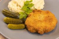 Golden breaded Dutch Schnitzel (Holandský Řízek) on a grey plate with mashed potato, gherkins and parsley.