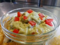 Glass bowl of Guacamole topped with creamy sauce and diced tomato, served with tortilla chips.