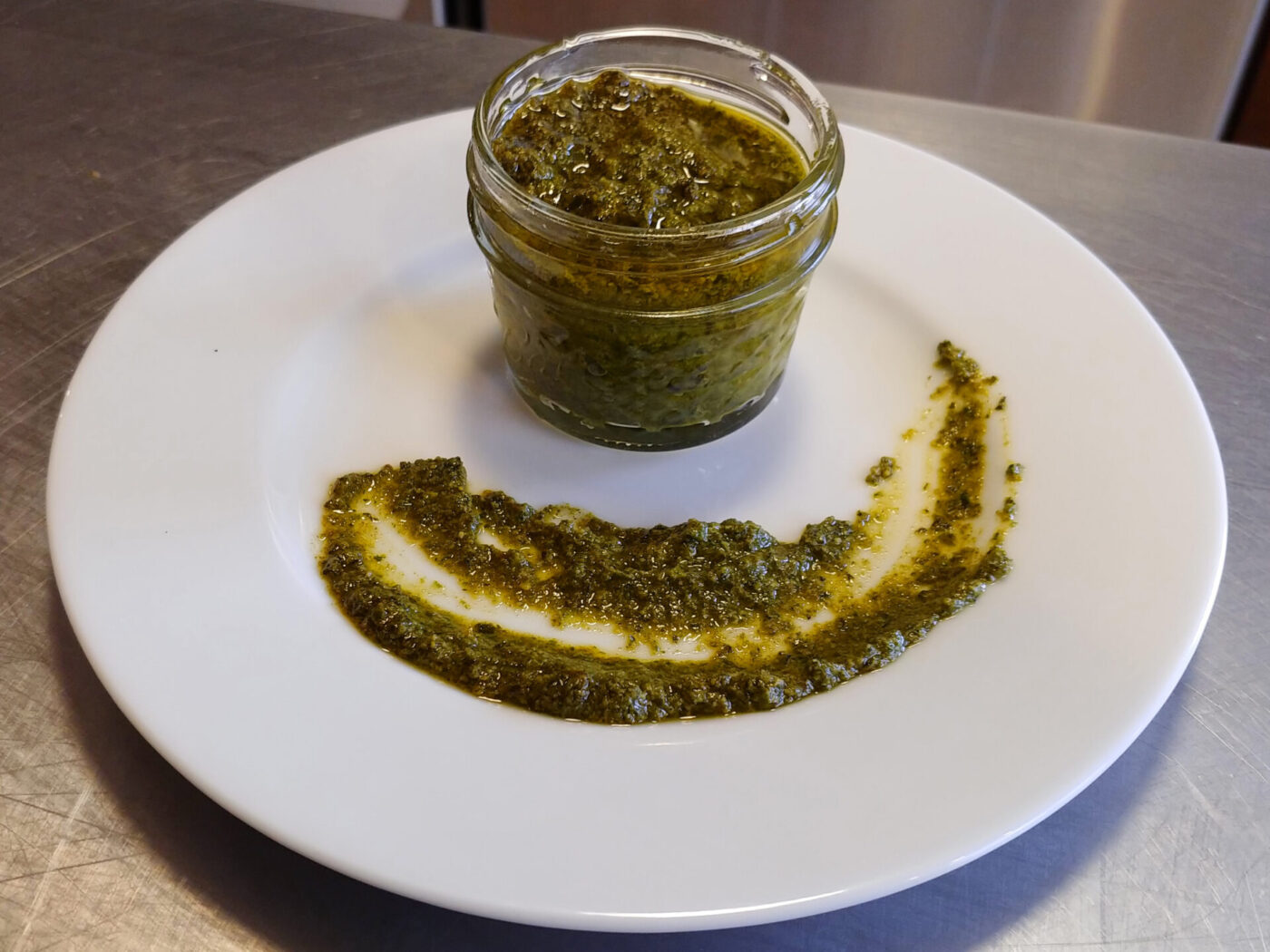 A glass jar of pesto beside a swirl of the same green sauce on a white plate.