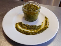A glass jar of pesto beside a swirl of the same green sauce on a white plate.
