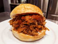 Pulled pork sandwich with red BBQ sauce on a soft bun, served on a white plate.