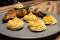 Seared Rosemary Chicken Breasts with green herb sauce, rosemary sprig, potato swirls, and bacon-wrapped asparagus.