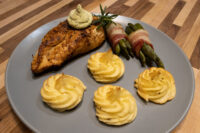 Rosemary Chicken Breasts with herb butter, duchess potatoes and bacon-wrapped green beans on a grey plate.