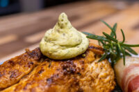 Seared Rosemary Chicken Breast topped with green herb butter, rosemary sprig and cured meat curl alongside.