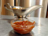 BBQ Sauce A glass bowl of chunky BBQ Sauce sits in front of a stainless steel gravy boat on a metal counter.