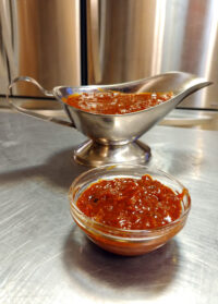 BBQ Sauce A silver gravy boat and a small glass bowl both filled with chunky BBQ sauce on a steel countertop.