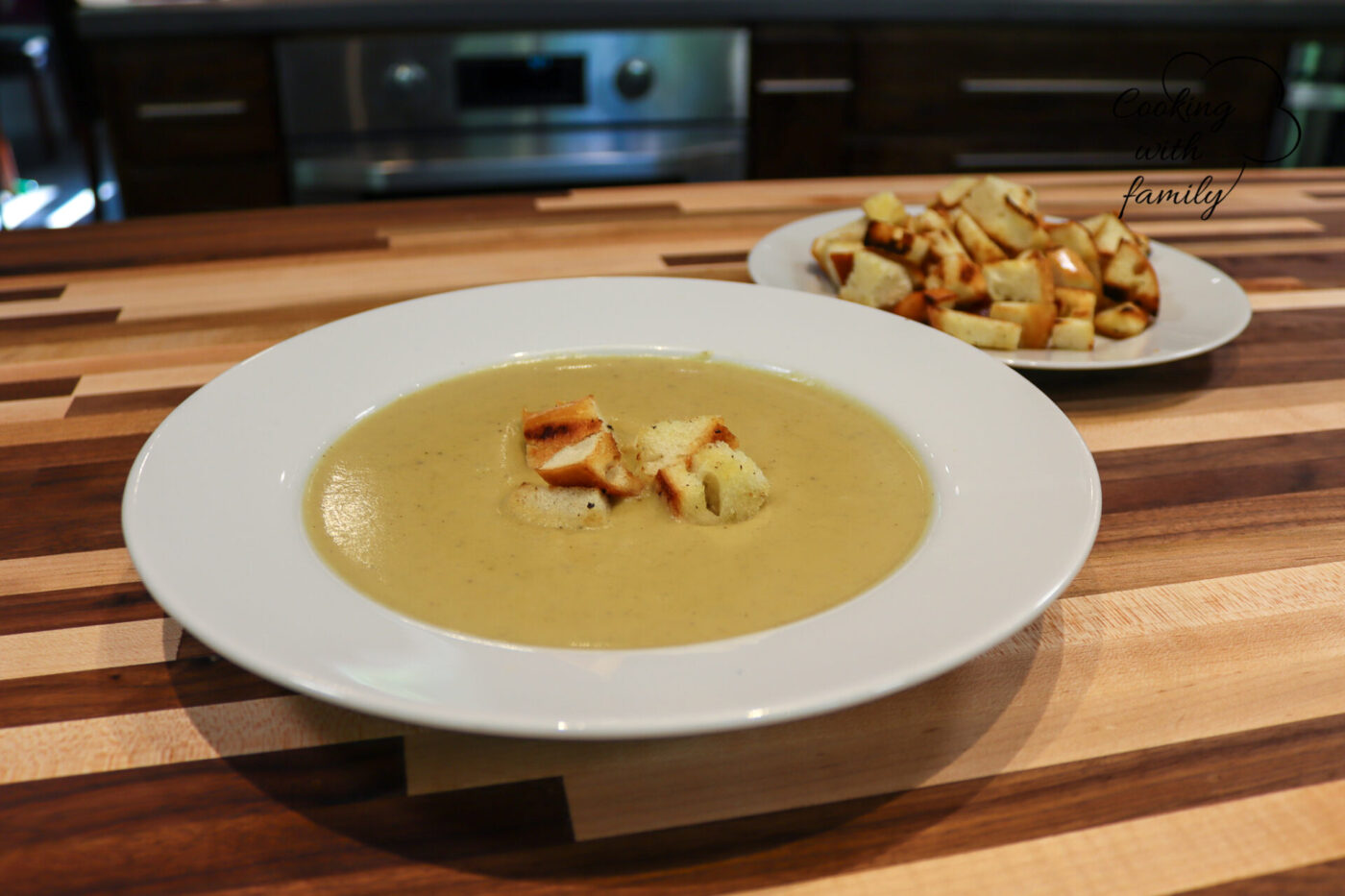 Bowl of creamy Broccoli Soup topped with croutons, extra croutons on a plate behind it on a wooden table.