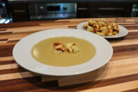 Bowl of creamy Broccoli Soup topped with croutons, extra croutons on a plate behind it on a wooden table.