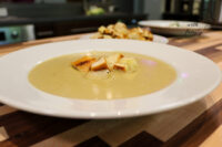 A white bowl of pale, creamy Broccoli Soup topped with golden croutons on a wooden table.