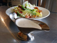 A metal gravy boat of Caesar Dressing (easy version) sits in front of a Caesar salad on a steel table.
