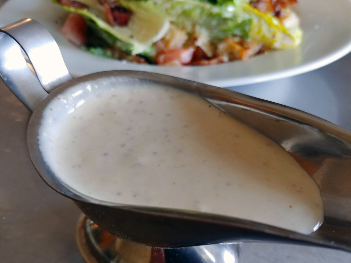 Creamy Caesar Dressing (easy version) pooled in a metal sauce boat, salad blurred behind it.