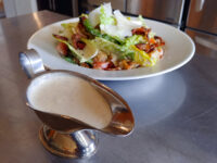 Creamy Caesar Dressing filling a metal gravy boat, set in front of a bacon and romaine salad.
