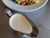 Creamy Caesar Dressing with flecks of seasoning pooled in a small metal creamer, salad plate blurred behind.