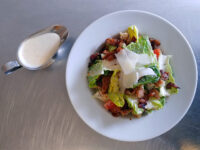 Caesar Salad with romaine, shaved parmesan, bacon, and tomato on a white plate, dressing in a metal pitcher nearby.