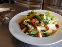 White plate of Caesar Salad with romaine leaves, croutons, shaved cheese, and diced tomato.