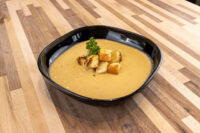 Cauliflower Soup Black square bowl of Cauliflower Soup topped with croutons and parsley, on a wooden table.