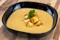 Cauliflower Soup Black bowl of Cauliflower Soup topped with toasted croutons and a parsley sprig on a wooden table.