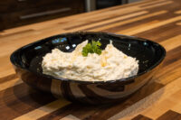 Cheese – Garlic Dip and Spread mounded in a dark oval bowl, topped with a parsley sprig, on a wooden counter.