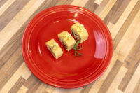 Three sliced rounds of Cheese Roulade with Goat Cheese & Raisins topped with piped cream, on a red plate with rosemary.