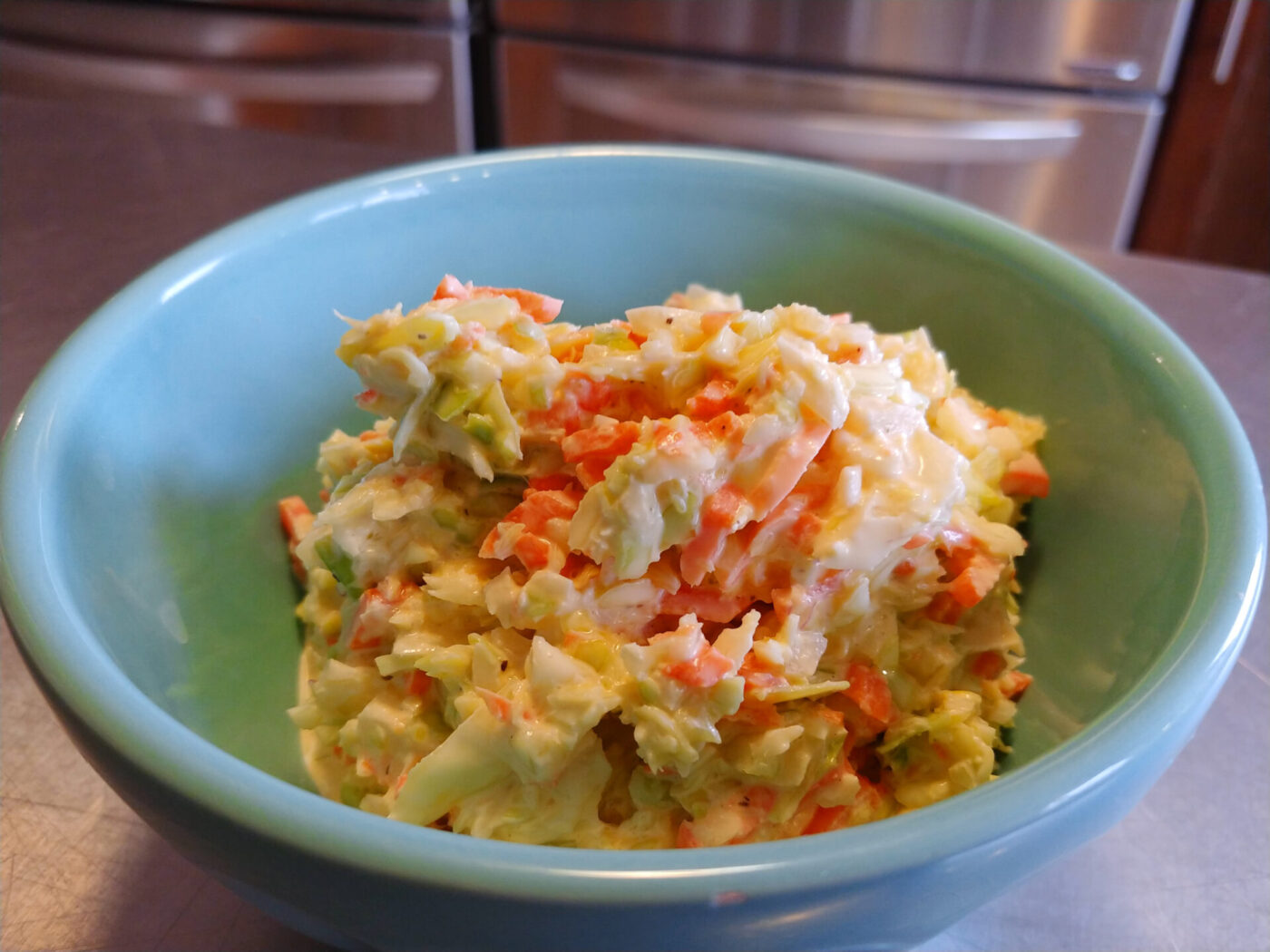 Turquoise bowl of Coleslaw with shredded cabbage and carrots in creamy dressing on a steel counter.
