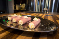 Five Creamy Ham Rolls piped with white filling and garnished with herbs, arranged on an oval metal tray on a striped wooden table