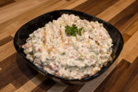 Black bowl of creamy Vlašský Salát Vlašák with peas and diced vegetables, garnished with parsley on a wooden table.
