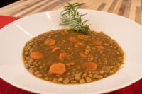 White bowl of Czech Lentil Soup Čočková Polévka with sliced carrots and a fresh herb sprig, on red cloth.