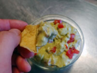 Hand dips a tortilla chip into a bowl of guacamole with diced tomato on a steel counter.