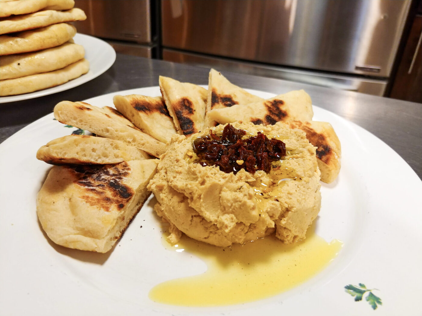 Hummus mounded on a plate, topped with a dark relish and olive oil, ringed by grilled flatbread wedges.