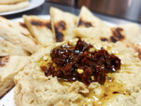 Bowl of hummus topped with chopped sun-dried tomatoes and olive oil, served with grilled pita triangles.