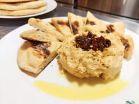 Round mound of hummus topped with dark relish and olive oil, ringed by grilled pita wedges on a white plate.
