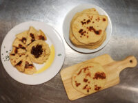 Bowl of hummus topped with a dark red garnish and olive oil, ringed by pita wedges, with extra flatbreads on the side.