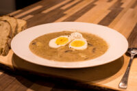 Kulajda Soup White bowl of Kulajda Soup with mushrooms, topped with two soft-boiled egg halves, served with bread.