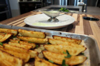 Pale green Mediterranean Aioli in a gravy boat beside roasted potato wedges on a baking tray.