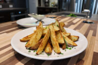 Golden Oven-Roasted Potato Wedges piled on a white plate, topped with chopped green chives, on a wooden counter.