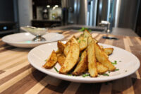 Plate of Oven-Roasted Potato Wedges topped with chives, with a small gravy boat of sauce behind it.