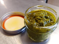 A glass jar filled with green pesto, its metal lid resting on a steel counter beside it.