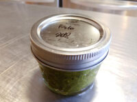 Small glass jar labeled 'Pesto 2oz' filled with green pesto, sitting on a steel countertop.