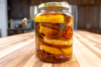 Glass jar of Pickled Brie Cheese Nakladany Hermelin wedges in oil with onion strands and herbs on a wood counter.