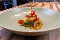 Round of Pickled Brie Cheese Nakladany Hermelin with onion rings, charred peppers, herbs, and oil on pale green plate