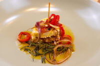 Wedge of Pickled Brie Cheese Nakladany Hermelin topped with red pepper and onion, skewered, on oiled rosemary sprigs.
