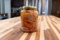 Glass jar of Pickled Sausage Utopenec with onion strands and red pepper in brine on a wood counter.