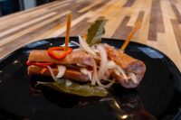Sliced Pickled Sausage Utopenec sausage links topped with onion and chili on a black plate.