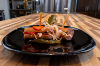 Pickled Sausage Utopenec: sliced sausages with pickled onions, red pepper strips, and bay leaves on a black plate.