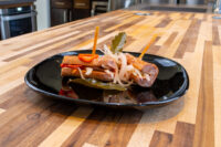 Pickled Sausage Utopenec with grilled sausage, onions, pepper strips, and bay leaf on a black plate.
