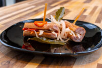 Two skewered rolled sausages of Pickled Sausage Utopenec with pickled onions, red pepper, and bay leaf on a black plate.