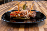 Two browned sausages of Pickled Sausage Utopenec topped with onion, pepper, and bay leaf on a black plate.